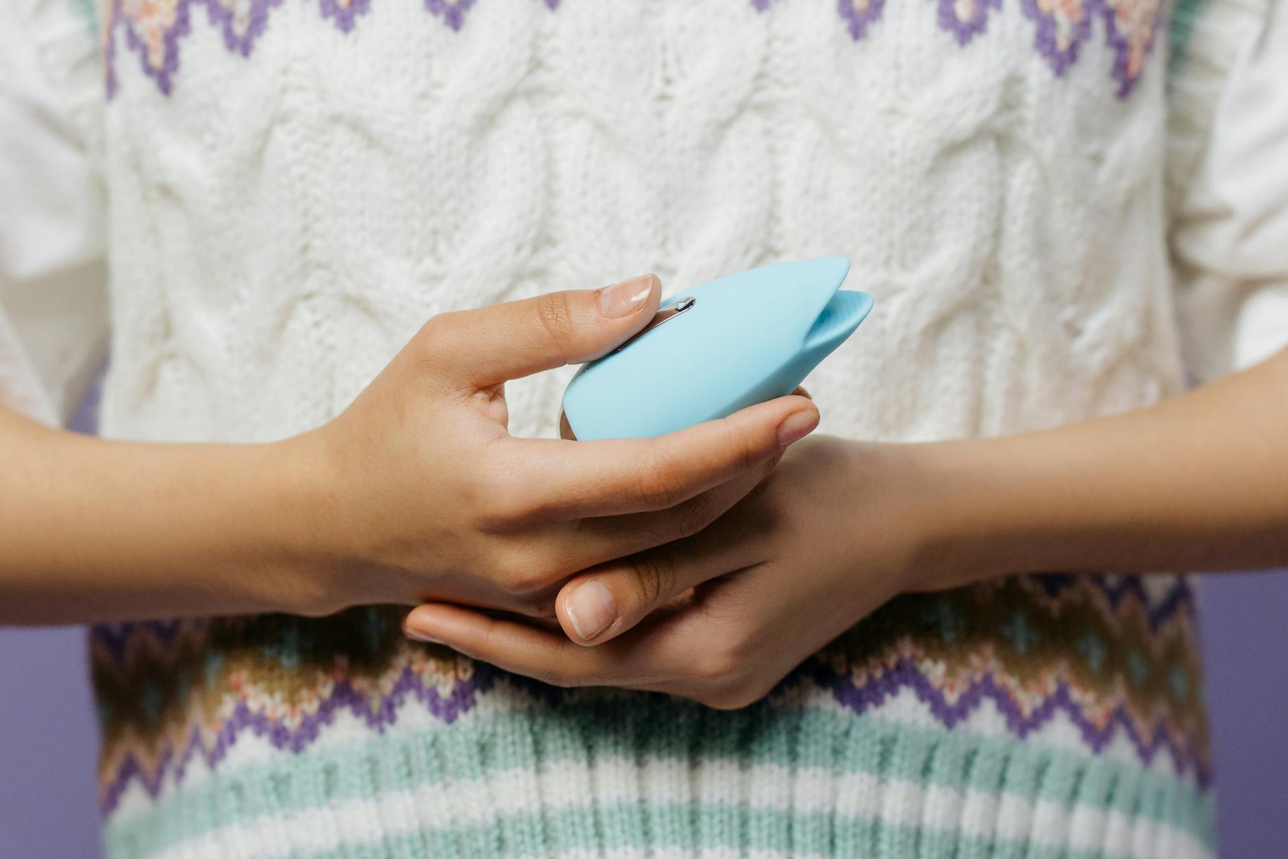 Close-up of hands holding a blue personal massager against a knitted sweater, highlighting intimate wellness tools.